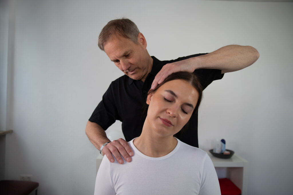 Ein Physiotherapeut behandelt den Nacken einer Patientin mit einer manuellen Mobilisation. Die Patientin sitzt entspannt aufrecht und neigt den Kopf zur Seite.